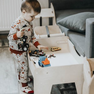 May include: A young child plays with toy trucks and cars on a white wooden table with a gray couch in the background.