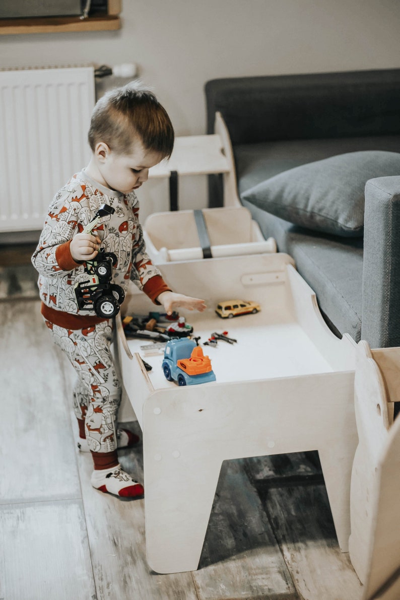 May include: A young child plays with toy trucks and cars on a white wooden table with a gray couch in the background.