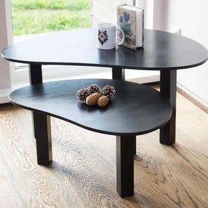May include: A black nesting coffee table set with two irregularly shaped tops. The top table has a mug and a book on it. The bottom table has pine cones on it.