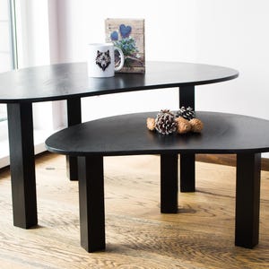 May include: Two black oval-shaped nesting coffee tables with black legs. The top table has a white mug with a wolf design on it and a small book with a blue and white floral design on it. The bottom table has a few pine cones on it.