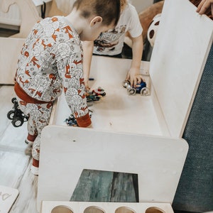 May include: Two children play with toy cars inside a white wooden playhouse with a removable lid. The playhouse has a built-in storage shelf with four round openings.