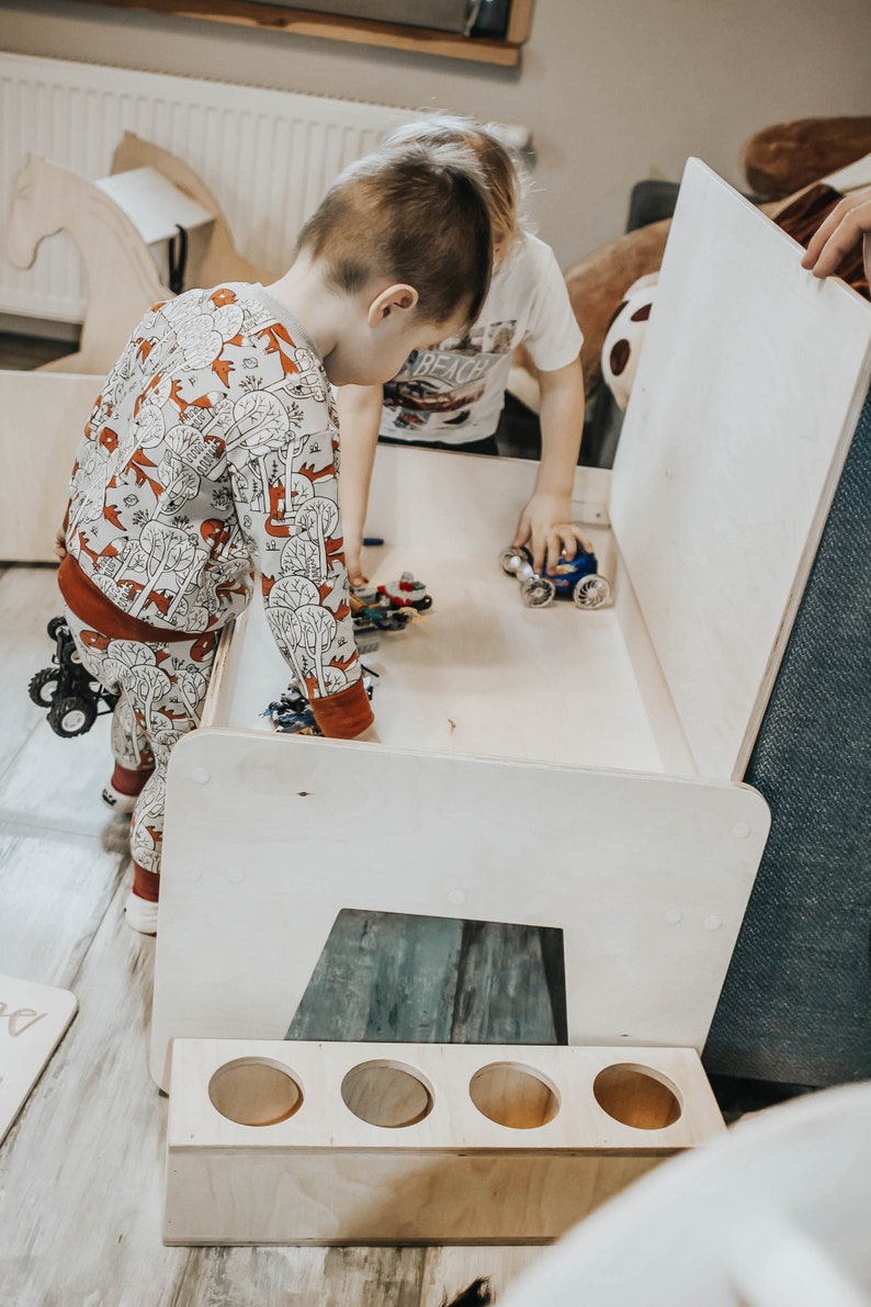 May include: Two children play with toy cars inside a white wooden playhouse with a removable lid. The playhouse has a built-in storage shelf with four round openings.