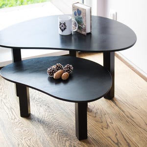 May include: Two black nesting coffee tables with a unique organic shape. The top table has a book and a white mug with a wolf design on it. The bottom table has a few pine cones on it.