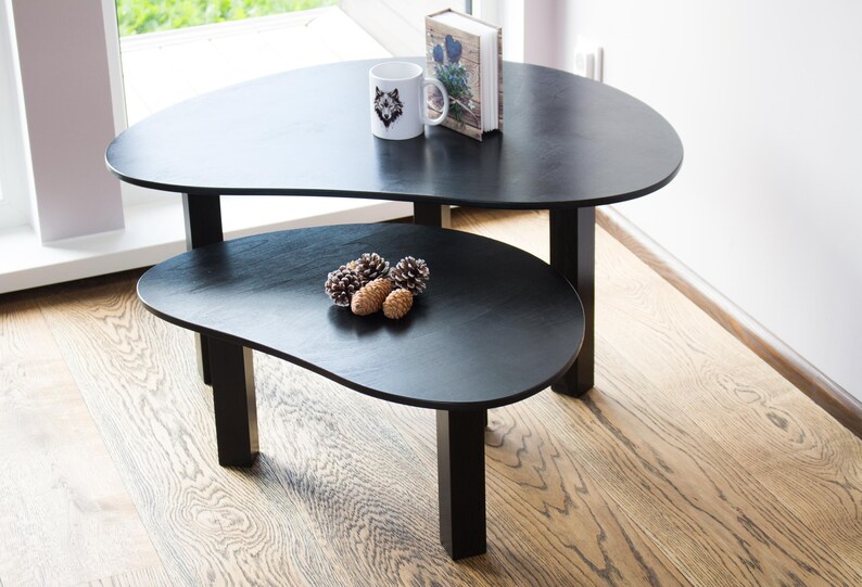 May include: Two black nesting coffee tables with a unique organic shape. The top table has a book and a white mug with a wolf design on it. The bottom table has a few pine cones on it.