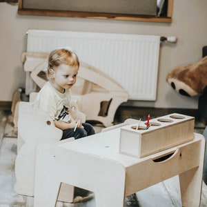 May include: A child sits at a wooden table and chair set, designed for kids. The table has a built-in storage compartment for crayons or other art supplies. The chair is shaped like a bear.