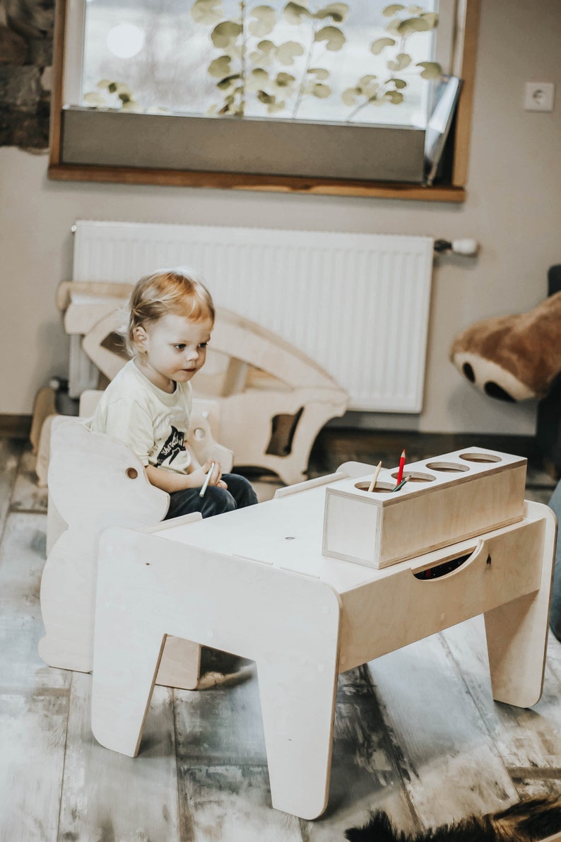 May include: A child sits at a wooden table and chair set, designed for kids. The table has a built-in storage compartment for crayons or other art supplies. The chair is shaped like a bear.