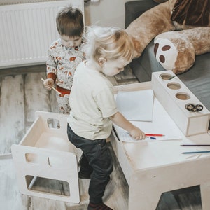 May include: Two children sitting at a wooden children's table and chairs. The table has a white surface and a built-in pencil holder. One child is drawing on the table with a red and blue crayon.