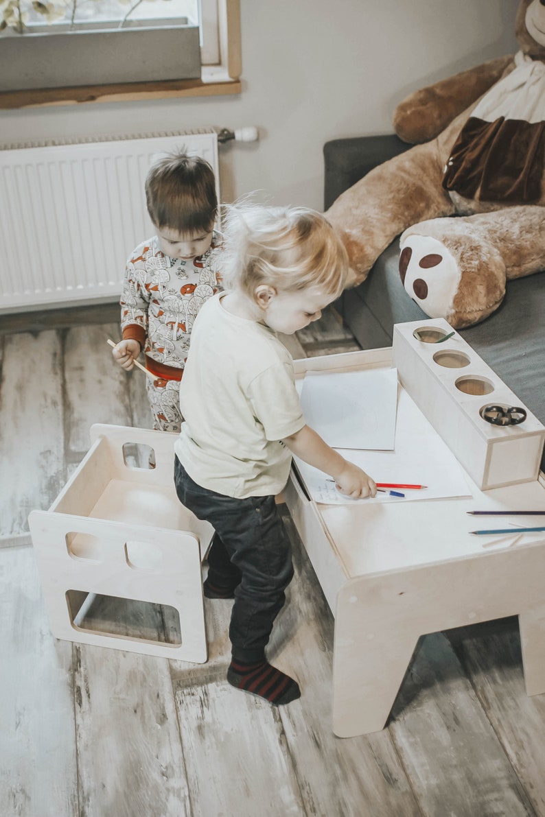 May include: Two children sitting at a wooden children's table and chairs. The table has a white surface and a built-in pencil holder. One child is drawing on the table with a red and blue crayon.