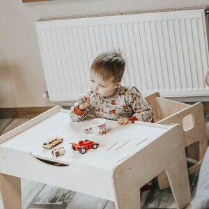 May include: A white wooden table with a child sitting at it. The table has a drawer and a small shelf. The child is holding a spoon and eating from a small cup. There are toy cars on the table.
