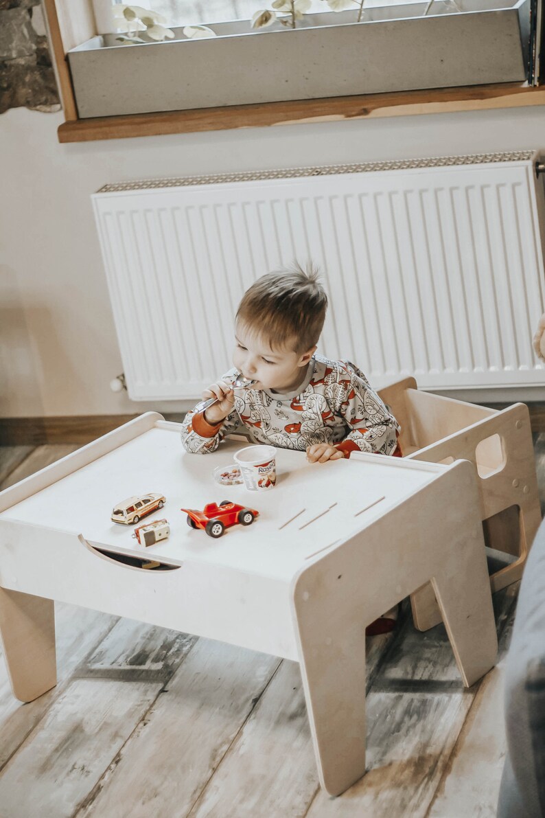 May include: A white wooden table with a child sitting at it. The table has a drawer and a small shelf. The child is holding a spoon and eating from a small cup. There are toy cars on the table.