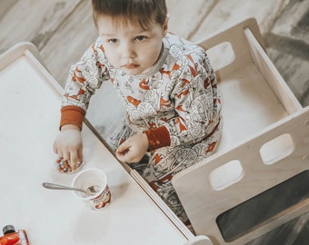 May include: A light-colored wooden learning tower with a child wearing a gray and red patterned pajama set. The child is seated at a table with a small red toy car, a yogurt cup, and a container of colorful candies.