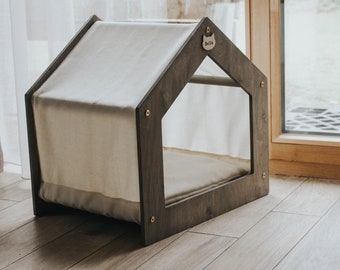Puede incluir: Una casa para mascotas de madera oscura con un techo de tela blanca y un cojín blanco en el interior. La casa tiene una pequeña ventana y una etiqueta con el nombre "Bella".