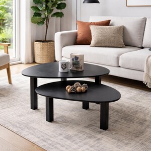 Two matching black coffee tables styled in a contemporary interior with a light-colored sofa and window in the background, decorated with a mug, a rustic wooden accent, and a cluster of pinecones.