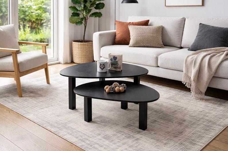 Two matching black coffee tables styled in a contemporary interior with a light-colored sofa and window in the background, decorated with a mug, a rustic wooden accent, and a cluster of pinecones.