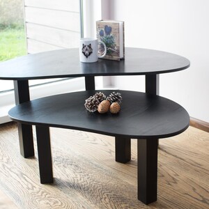 May include: Two black nesting tables with a unique organic shape. The top table has a white mug with a wolf design and a book on top. The bottom table has three pine cones on top.