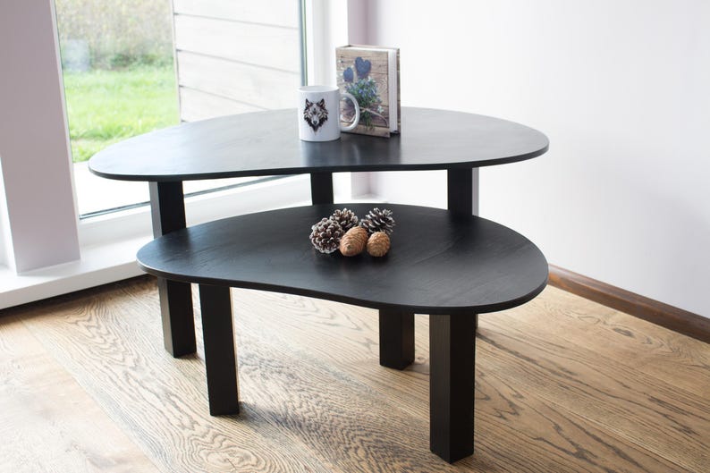 May include: Two black nesting tables with a unique organic shape. The top table has a white mug with a wolf design and a book on top. The bottom table has three pine cones on top.