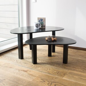 May include: Two black oval-shaped coffee tables with black metal legs. The top table has a small white mug with a wolf design on it and a small book with a floral design on it. The bottom table has a small pile of pine cones on it.