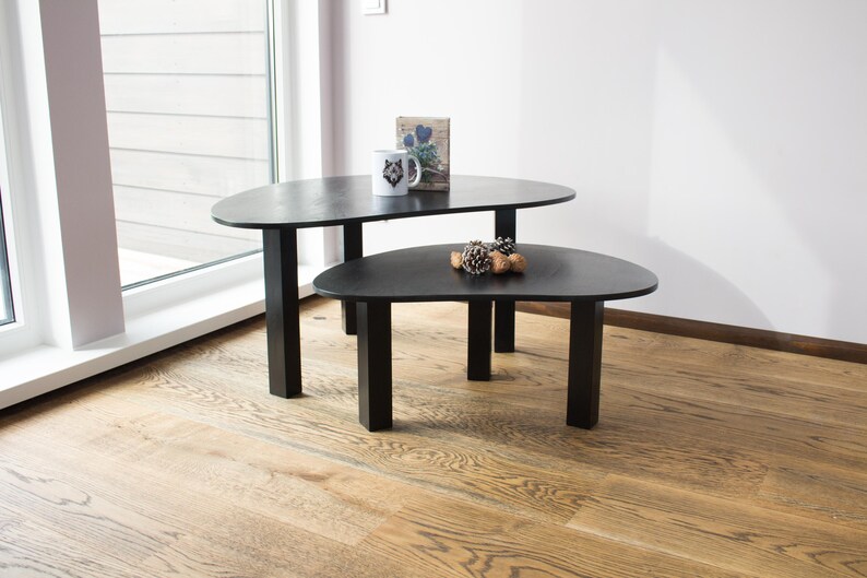 May include: Two black oval-shaped coffee tables with black metal legs. The top table has a small white mug with a wolf design on it and a small book with a floral design on it. The bottom table has a small pile of pine cones on it.