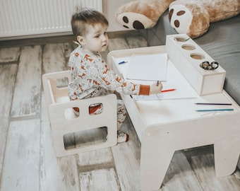 May include: A child sits at a white wooden table and chair set, drawing on a piece of paper with a red crayon. The table has a built-in pencil holder and a small shelf for storing supplies.