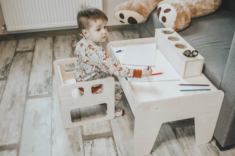 May include: A child sits at a white wooden table and chair set, drawing on a piece of paper with a red crayon. The table has a built-in pencil holder and a small shelf for storing supplies.