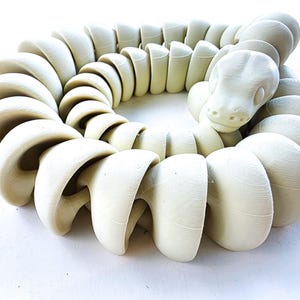 Puede incluir: Una escultura de serpiente articulada de color crema. La serpiente está enrollada, con una cabeza detallada y un cuerpo segmentado. La escultura está hecha de un material sólido y tiene un acabado liso y mate. El diseño es moderno y artístico.
