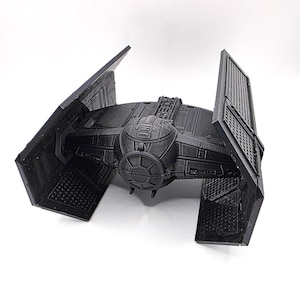 Pode incluir: Um modelo preto impresso em 3D de uma nave espacial TIE fighter. O modelo apresenta painéis detalhados, um cockpit esférico central e dois grandes painéis solares angulados. A nave espacial é um item popular da franquia Star Wars.