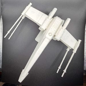 May include: A white model of an X-wing starfighter, a spacecraft from the Star Wars franchise. The model has four wings, two on each side, and four engines. The spacecraft is white and appears to be made of plastic.