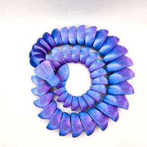 Puede incluir: Una escultura en espiral compuesta por numerosas piezas semicirculares. Las piezas transicionan en color de morado oscuro a azul claro, con un patrón de vetas de madera visible en cada una. La escultura está sobre un fondo blanco.