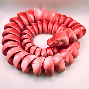 Peut inclure: Un jouet serpent segmenté de couleur corail enroulé en cercle. La tête du serpent est relevée, avec un œil détaillé. Le corps est fait de pièces arrondies et imbriquées, créant un design articulé et flexible. Le jouet est probablement imprimé en 3D.