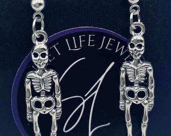 Puede incluir: Pendientes de esqueleto plateados. Cada pendiente presenta una pequeña calavera y un colgante de esqueleto completo. Los pendientes cuelgan de un pequeño perno de bola. El fondo es azul oscuro con el texto "Handmade with love".