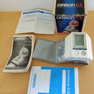 Puede incluir: Un tensi&oacute;metro de mu&ntilde;eca OMRON RX blanco con pantalla digital, botones y un manguito gris. El dispositivo viene con su caja original, manuales de instrucciones y una tarjeta de garant&iacute;a. La caja es azul y blanca.
