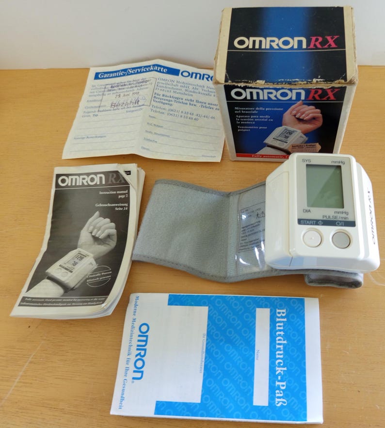 Puede incluir: Un tensi&oacute;metro de mu&ntilde;eca OMRON RX blanco con pantalla digital, botones y un manguito gris. El dispositivo viene con su caja original, manuales de instrucciones y una tarjeta de garant&iacute;a. La caja es azul y blanca.