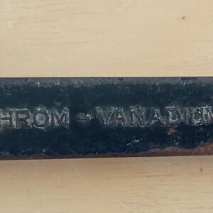 Peut inclure: Gros plan d'une clé noire avec le texte "CHROM - VANADIEM" en relief sur sa surface. L'outil montre des signes d'usure, avec un aspect légèrement rouillé. L'arrière-plan est une surface en bois brun clair.
