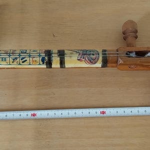 Può includere: Uno strumento a corde in legno fatto a mano con una cassa armonica rotonda e un lungo manico decorato. Lo strumento presenta intagli intricati e dettagli dipinti, con un'estremità nera a forma di corno. Un righello è posto accanto allo strumento.