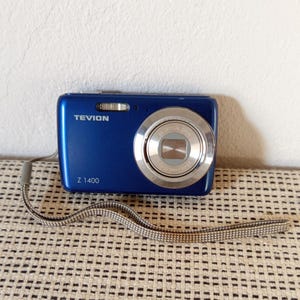 Op de afbeelding: Een blauwe Tevion Z 1400 digitale camera met een zilverkleurige lensbehuizing met de tekst 5X optical zoom. De camera heeft een zilverkleurige lensring en een grijs geweven polsbandje aan de zijkant, met de merknaam en het modelnummer zichtbaar.