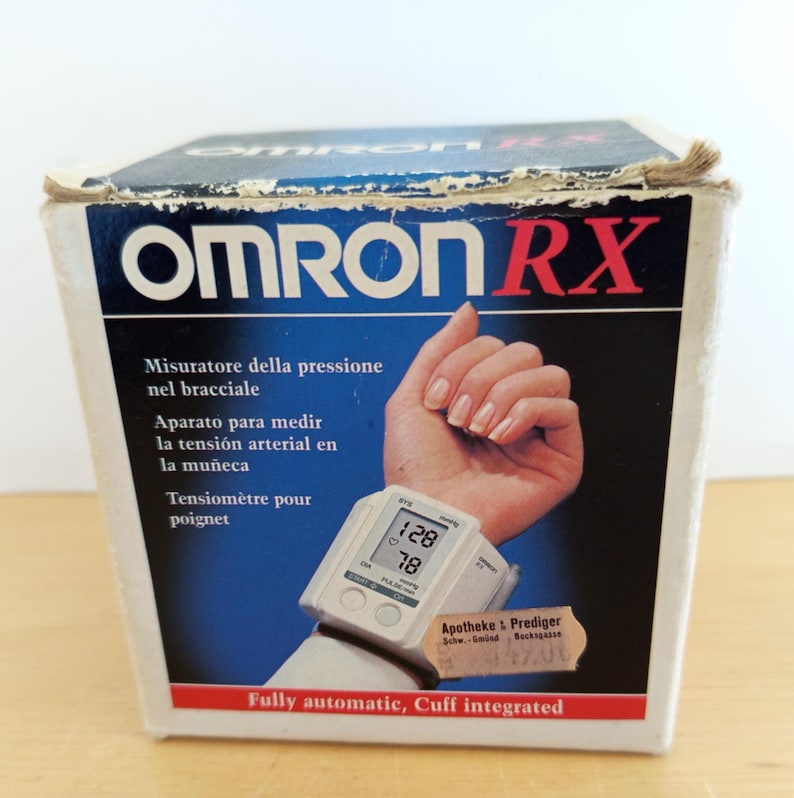 Puede incluir: Caja blanca y azul con un tensi&oacute;metro de mu&ntilde;eca OMRON RX. El monitor muestra lecturas de 128/78. La caja incluye texto en varios idiomas, incluido el espa&ntilde;ol. Totalmente autom&aacute;tico, manguito integrado.