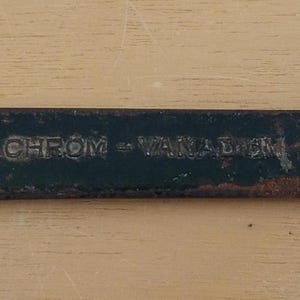 Könnte beinhalten: Ein doppelseitiger Schraubenschlüssel mit dunkelgrünem Körper und rostigen, gebogenen Enden. Der Schraubenschlüssel hat schwer lesbaren Text. Dieses Werkzeug dient zum Anziehen oder Lösen von Muttern und Bolzen.