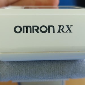 Puede incluir: Un dispositivo m&eacute;dico blanco con el texto "OMRON RX" en negro. El dispositivo descansa sobre una superficie acolchada gris. La imagen es un primer plano, destacando el dise&ntilde;o y la marca del producto.