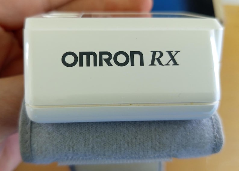 Puede incluir: Un dispositivo m&eacute;dico blanco con el texto "OMRON RX" en negro. El dispositivo descansa sobre una superficie acolchada gris. La imagen es un primer plano, destacando el dise&ntilde;o y la marca del producto.