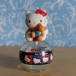 Puede incluir: Un frasco de eau de toilette de Hello Kitty. El frasco redondo y transparente tiene una banda negra con imágenes de Hello Kitty. Una figura blanca de Hello Kitty con un lazo rojo y un pequeño oso de peluche se encuentra en la parte superior.