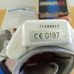 Puede incluir: Un monitor de presi&oacute;n arterial blanco con un manguito gris. El monitor tiene los n&uacute;meros "71209517" y "CE 0197" impresos. La caja en el fondo dice "OMRON RX".