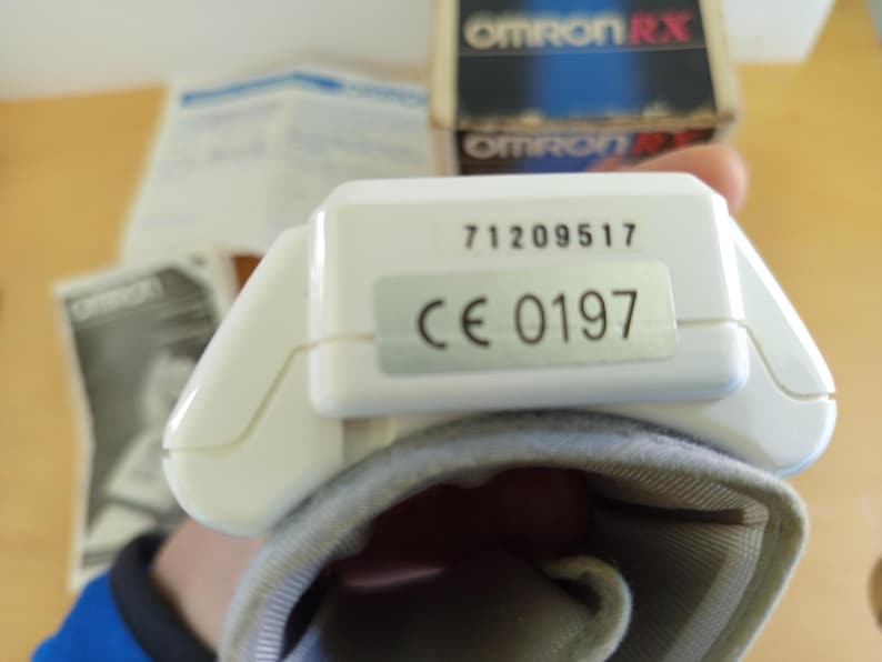 Puede incluir: Un monitor de presi&oacute;n arterial blanco con un manguito gris. El monitor tiene los n&uacute;meros "71209517" y "CE 0197" impresos. La caja en el fondo dice "OMRON RX".