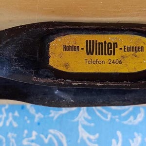 Op de afbeelding: Een vintage zwart metalen gereedschap met een gebogen handvat en een plat, rechthoekig uiteinde. Een geel label met zwarte tekst met de tekst "Kohlen - Winter - Ebingen, Telefon 2406" is op het gereedschap bevestigd. Het gereedschap ligt op een houten oppervlak.