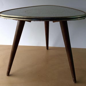Può includere: Un tavolino triangolare vintage con tre gambe in legno scuro. Il piano del tavolo ha una superficie grigio chiaro strutturata con un bordo dorato e nero. Il tavolo misura circa 61 cm di larghezza e 51 cm di profondità.