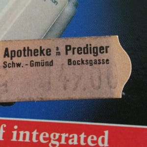 Puede incluir: Primer plano de una etiqueta de color beige con texto negro y bordes dentados. El texto dice "Apotheke am Prediger" y "Schw.-Gm&uuml;nd Bocksgasse". La etiqueta oscurece parcialmente un fondo azul y blanco con texto rojo en la parte inferior.