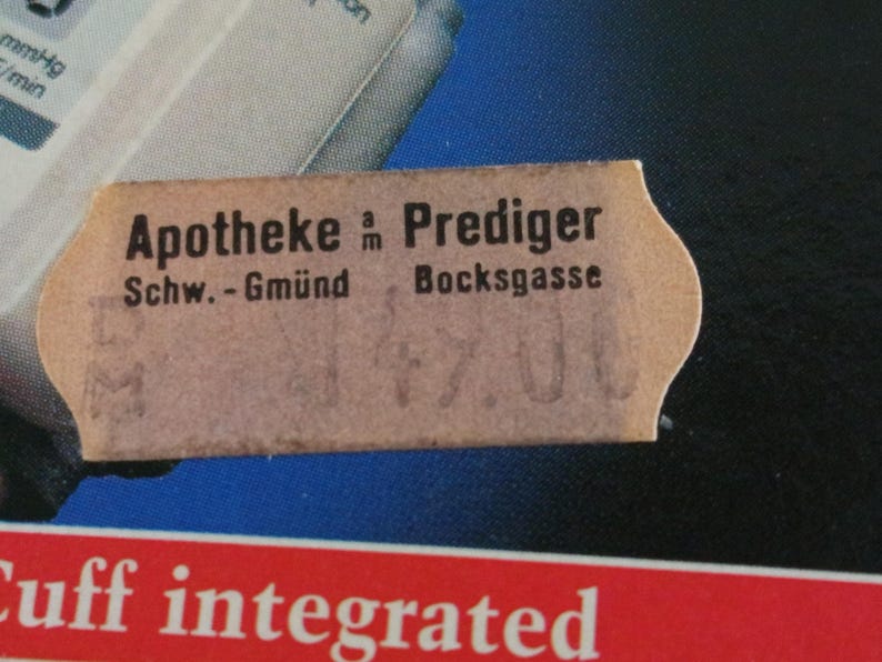 Puede incluir: Primer plano de una etiqueta de color beige con texto negro y bordes dentados. El texto dice "Apotheke am Prediger" y "Schw.-Gm&uuml;nd Bocksgasse". La etiqueta oscurece parcialmente un fondo azul y blanco con texto rojo en la parte inferior.