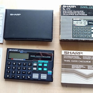 May include: A collection of vintage Sharp EL-470 Travel Clock Calculators. The image shows a black calculator, a black case, a box, and instruction manuals. The calculator has a digital display and a keypad with various function keys.