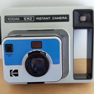 Puede incluir: Una cámara instantánea Kodak EK2 vintage. La cámara es principalmente gris claro con un panel superior negro y un panel frontal azul y negro. La cámara tiene una lente, un visor y el texto "KODAK EK2 INSTANT CAMERA".