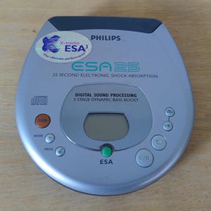 Può includere: Un lettore CD portatile Philips ESA25 argentato con parte superiore grigio scuro. Include un display digitale, pulsanti di controllo e il testo "ESA 25" e "Digital Sound Processing". Un adesivo olografico recita "X-treme ESA³".