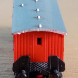 May include: A miniature red train caboose with a silver roof and black undercarriage. The caboose has a small rectangular window and several silver rivets along the roofline. This model train car is designed for a model railroad.
